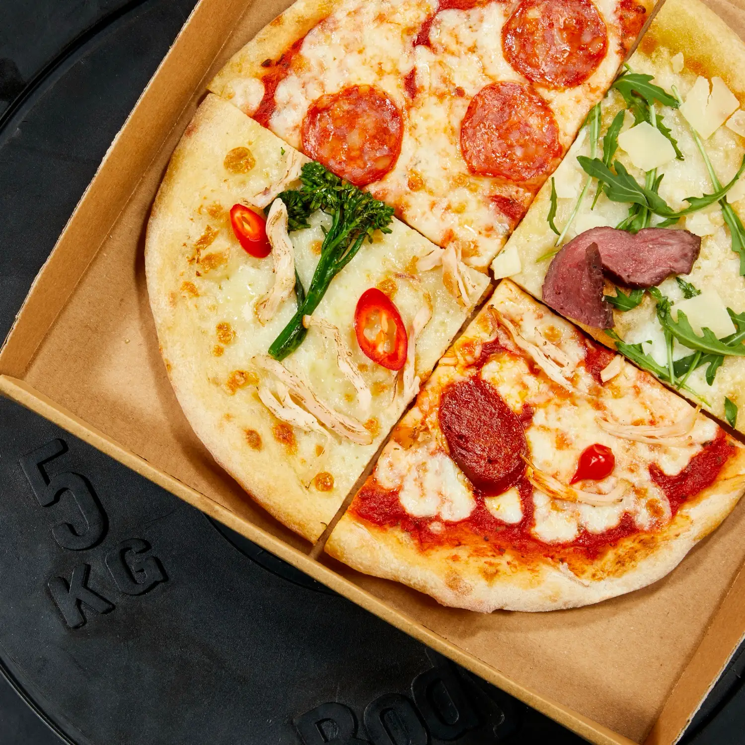 A Protein Pizza Co. box resting on a 5kg weight plate, the pizza cut into quarters with different toppings