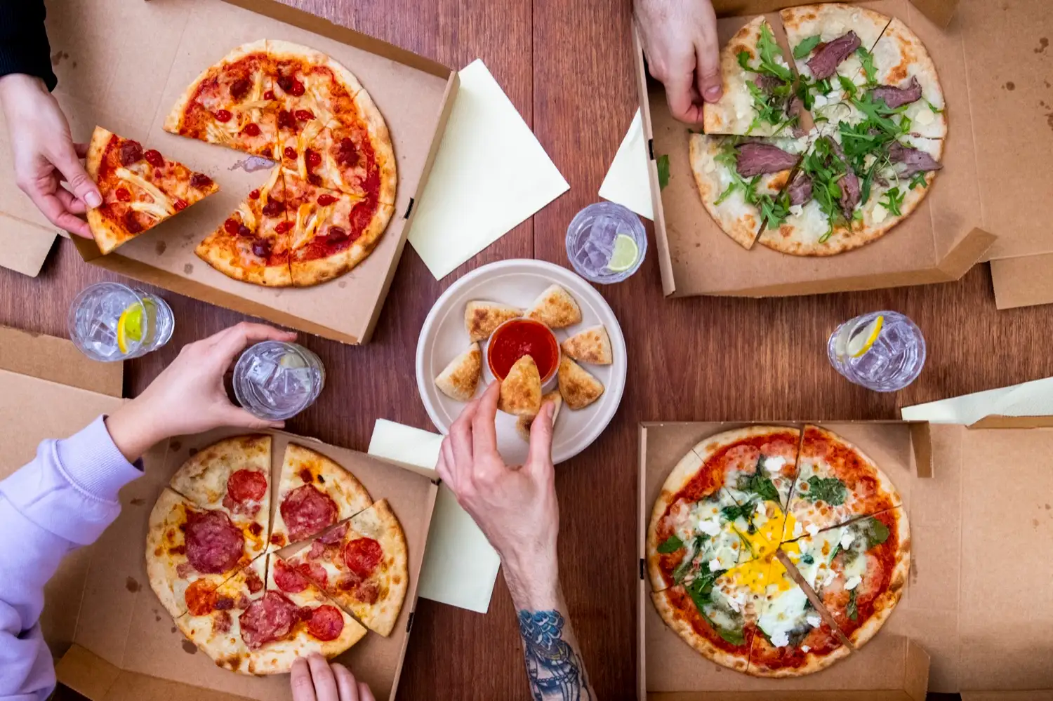 Four Protein Pizza Co. pizzas open in their boxes on a table, with several people reaching in to share and a plate of dough balls in the middle